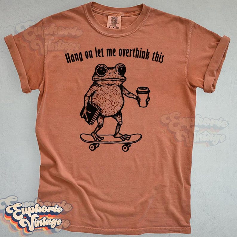 Puede incluir: Una camiseta de manga corta color terracota con un gr&aacute;fico negro de una rana en un monopat&iacute;n sosteniendo un libro y una taza de caf&eacute;. El texto sobre la rana dice, Hang on let me overthink this. La camiseta tiene un cuello redondo cl&aacute;sico.