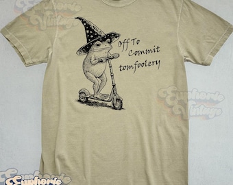 May include: Tan-colored t-shirt featuring a whimsical illustration of a frog wearing a wizard hat and riding a scooter. The text "Off To Commit Tomfoolery" is printed on the shirt. The shirt has a classic crew neck and short sleeves.