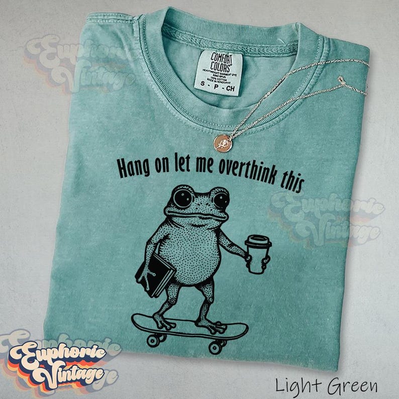 Puede incluir: Una camiseta verde claro con un gr&aacute;fico negro de una rana en un monopat&iacute;n sosteniendo un libro y una taza de caf&eacute;. El texto sobre la rana dice: "Hang on let me overthink this". La camiseta tiene cuello redondo y una etiqueta de la marca Comfort Colors.