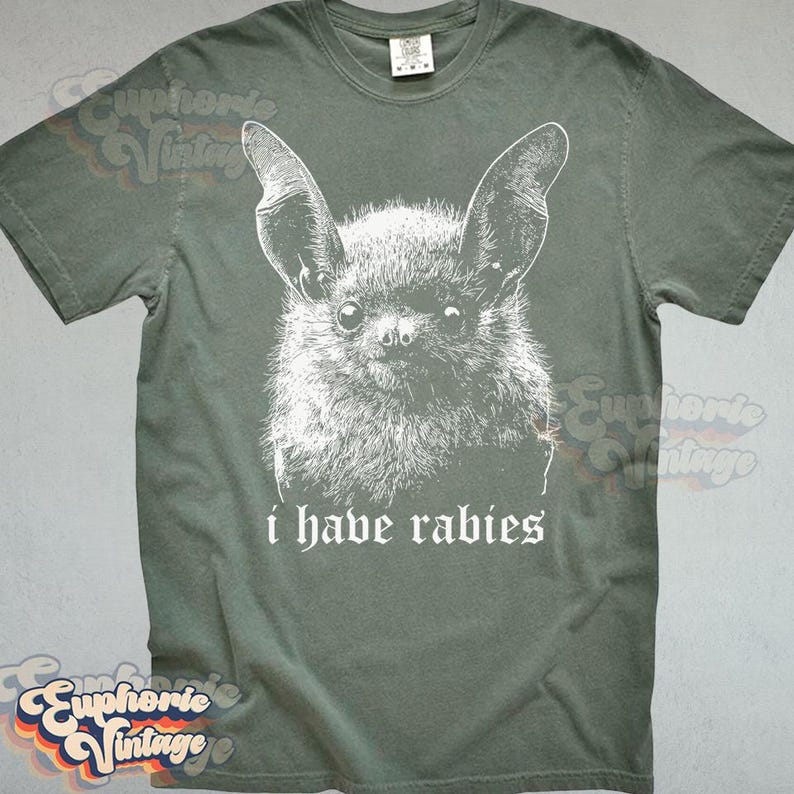 May include: A sage green t-shirt featuring a white illustration of a bat's face. Below the bat, the text reads "i have rabies" in a serif font. The shirt has a crew neck and short sleeves.