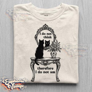 May include: A cream-colored t-shirt featuring a black graphic print of a cat sitting on a table in front of an ornate mirror. The text reads, I do not think therefore I do not am. The design includes a vase of flowers on the table next to the mirror.