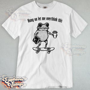 Puede incluir: Una camiseta blanca de manga corta con una ilustraci&oacute;n negra estilo tinta de una rana sobre un monopat&iacute;n, sosteniendo un libro y un vaso de caf&eacute;. Sobre la rana, el texto dice Hang on let me overthink this en una fuente negra sencilla.