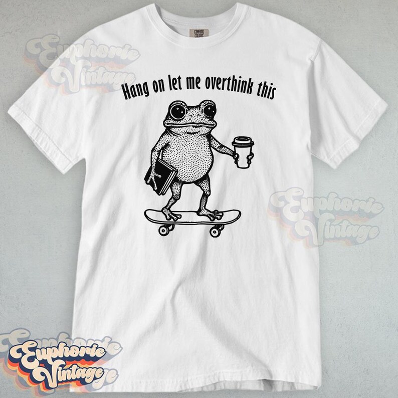 Puede incluir: Una camiseta blanca de manga corta con una ilustraci&oacute;n negra estilo tinta de una rana sobre un monopat&iacute;n, sosteniendo un libro y un vaso de caf&eacute;. Sobre la rana, el texto dice Hang on let me overthink this en una fuente negra sencilla.