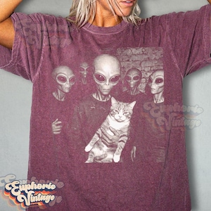 May include: Burgundy t-shirt featuring a graphic of several aliens holding a cat. The aliens have large black eyes and gray skin. The cat is gray and white. The shirt has the words "Euphorie Vintage" in a retro font.