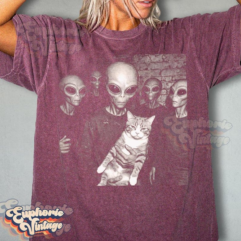 May include: Burgundy t-shirt featuring a graphic of several aliens holding a cat. The aliens have large black eyes and gray skin. The cat is gray and white. The shirt has the words "Euphorie Vintage" in a retro font.