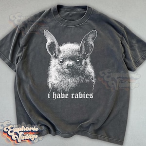 May include: Dark grey t-shirt featuring a detailed white illustration of a bat's face. Below the image, the text reads "i have rabies" in a gothic font. The shirt has a vintage, worn look.