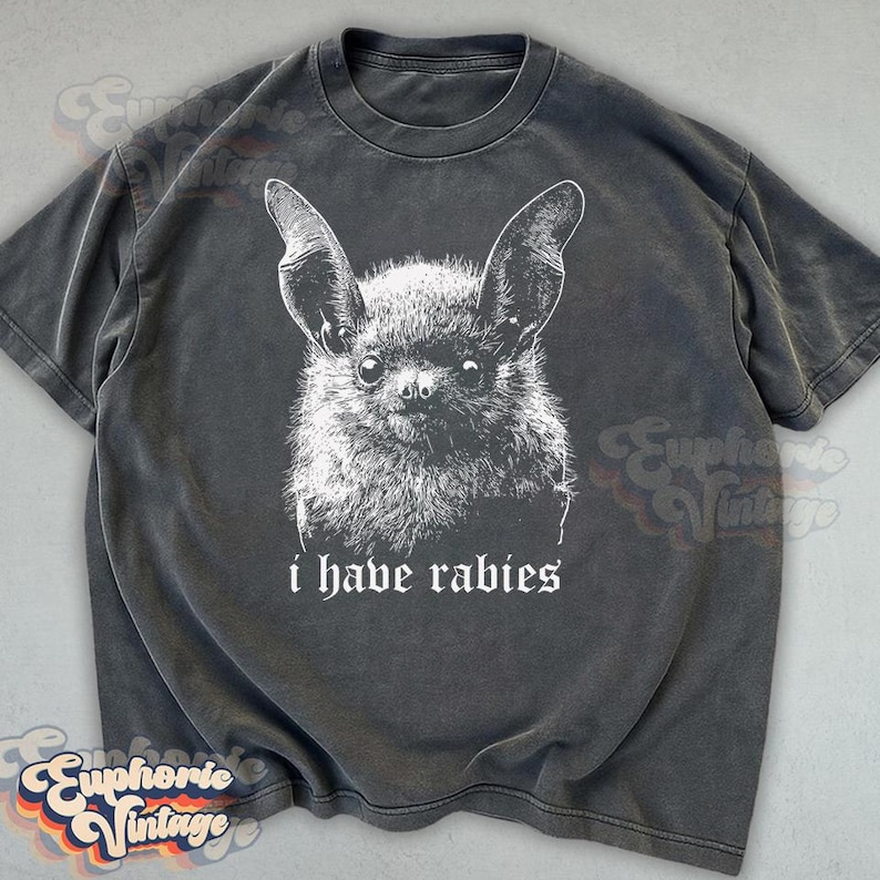 May include: Dark grey t-shirt featuring a detailed white illustration of a bat's face. Below the image, the text reads "i have rabies" in a gothic font. The shirt has a vintage, worn look.