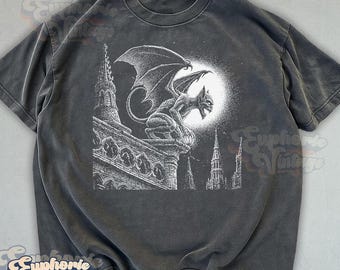 May include: A charcoal gray short-sleeve t-shirt featuring a white graphic print of a winged gargoyle perched on a gothic cathedral rooftop against a full moon. The design includes intricate architectural details and a dark, moody aesthetic.