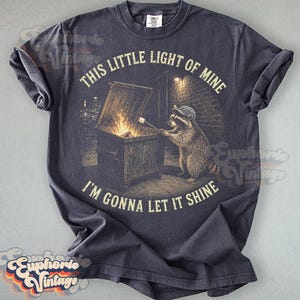 May include: A charcoal gray t-shirt featuring a graphic of a raccoon wearing a cap, standing by a dumpster fire and roasting a marshmallow on a stick. The text "THIS LITTLE LIGHT OF MINE" and "I'M GONNA LET IT SHINE" is printed in a circular arc.