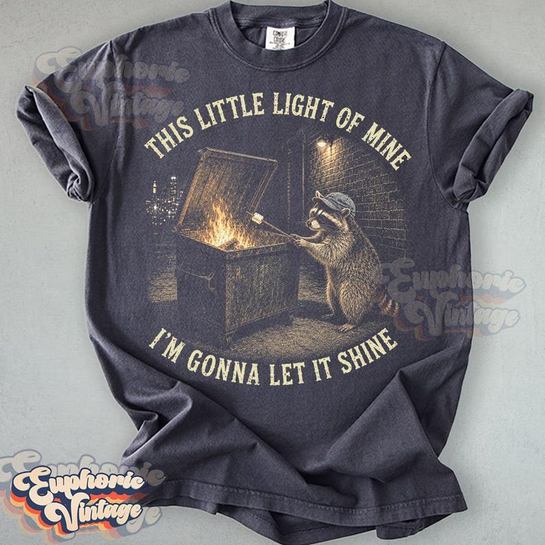 May include: A charcoal gray t-shirt featuring a graphic of a raccoon wearing a cap, standing by a dumpster fire and roasting a marshmallow on a stick. The text "THIS LITTLE LIGHT OF MINE" and "I'M GONNA LET IT SHINE" is printed in a circular arc.