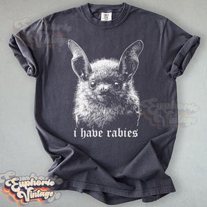 May include: Dark gray t-shirt featuring a detailed white illustration of a bat's face. The text "i have rabies" is printed below the bat. The shirt has rolled-up sleeves and a vintage aesthetic.