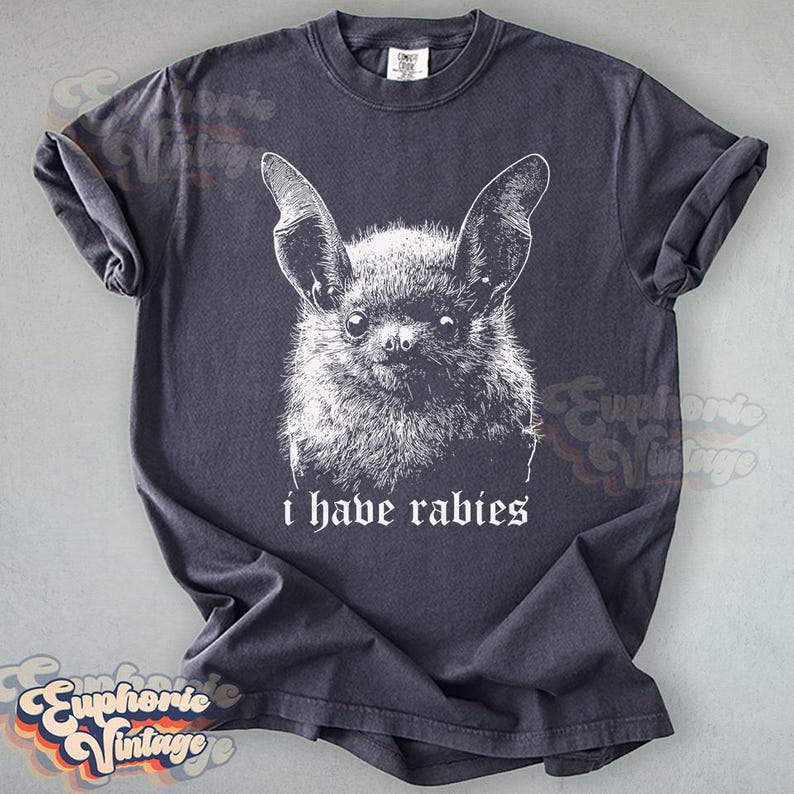 May include: Dark gray t-shirt featuring a detailed white illustration of a bat's face. The text "i have rabies" is printed below the bat. The shirt has rolled-up sleeves and a vintage aesthetic.