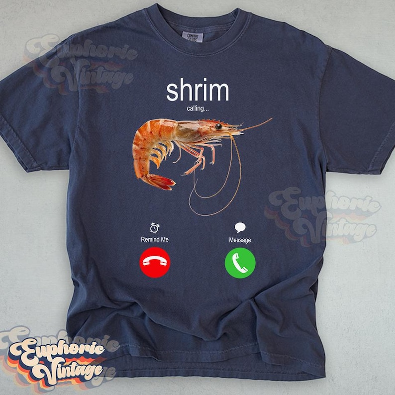 Op de afbeelding: Marineblauw t-shirt met een garnalenafbeelding en de tekst "shrimp calling...". Het ontwerp bevat een realistische garnalenillustratie en telefooninterface-pictogrammen voor "Remind Me" en "Message".