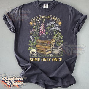 May include: A charcoal gray t-shirt featuring a graphic print of a black cat resting on a stack of books labeled Herbarium, Alchemia Herbalis, and Liber Mortis. The design includes a human skull, a mortar and pestle, flowers, and the text ALL PLANTS ARE EDIBLE SOME ONLY ONCE.