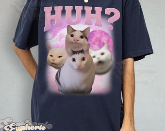 May include: Navy blue t-shirt featuring a graphic of four cats with surprised expressions and the word "HUH?" in pink. The design includes a pink moon and clouds. The shirt has a vintage style.