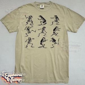 May include: A tan-colored t-shirt featuring a black graphic of nine frog skeletons in various poses. The shirt has a crew neck and short sleeves. The design is centered on the front of the shirt.
