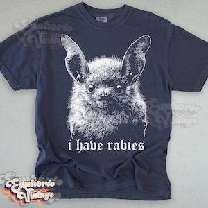 May include: Navy blue t-shirt featuring a detailed white illustration of a bat's face. The text "i have rabies" is printed below the bat. The shirt has a vintage aesthetic.