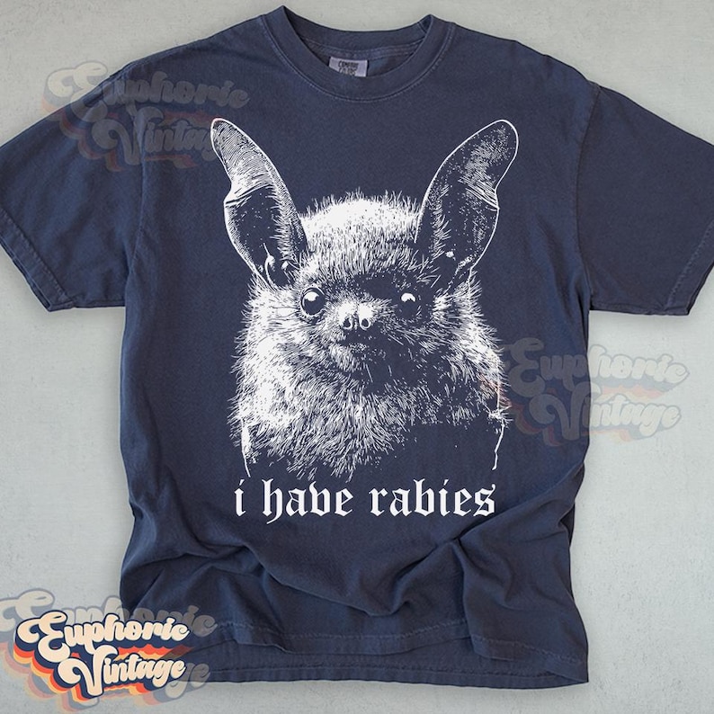 May include: Navy blue t-shirt featuring a detailed white illustration of a bat's face. The text "i have rabies" is printed below the bat. The shirt has a vintage aesthetic.