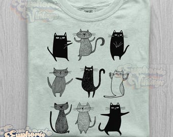 May include: A light mint green t-shirt featuring a graphic print of nine stylized cats in various poses. The cats are rendered in black and grey ink with distinct line work and textures. The shirt has a crew neckline and a visible interior neck label.