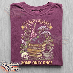 May include: A maroon t-shirt featuring a graphic print of a black cat sitting on a stack of books labeled Herbarium, Alchemia Herbalis, and Liber Mortis, surrounded by wildflowers, a skull, and a mortar and pestle with the text All Plants Are Edible Some Only Once.