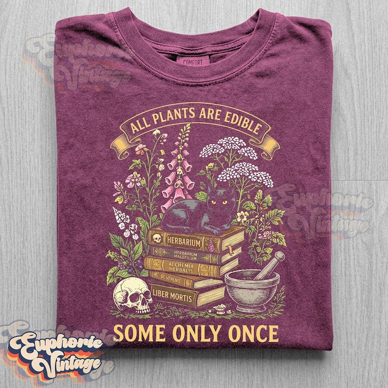 May include: A maroon t-shirt featuring a graphic print of a black cat sitting on a stack of books labeled Herbarium, Alchemia Herbalis, and Liber Mortis, surrounded by wildflowers, a skull, and a mortar and pestle with the text All Plants Are Edible Some Only Once.