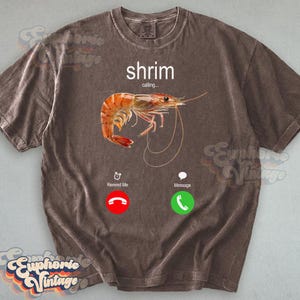Op de afbeelding: Bruin t-shirt met een afbeelding van een garnaal en de woorden "shrimp calling..." in het wit. Het ontwerp bevat een rood telefoonpictogram met "Remind Me" en een groen telefoonpictogram met "Message".