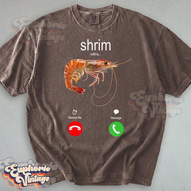 Op de afbeelding: Bruin t-shirt met een afbeelding van een garnaal en de woorden "shrimp calling..." in het wit. Het ontwerp bevat een rood telefoonpictogram met "Remind Me" en een groen telefoonpictogram met "Message".