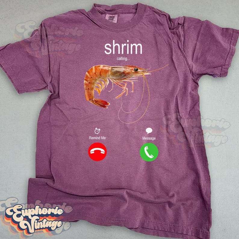 Op de afbeelding: Een paars T-shirt met de tekst "shrimp calling..." boven een realistische afbeelding van een gekookte garnaal. Onder de garnaal staan twee telefooniconen: "Remind Me" en "Message". Het shirt heeft een vintage stijl.