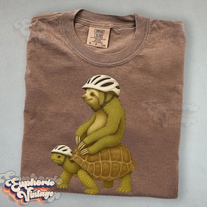 May include: A brown Comfort Colors t-shirt featuring a whimsical illustration of a sloth riding a turtle, both wearing white helmets. The sloth is olive green, and the turtle is brown. The shirt has a classic crew neck and a small tag.