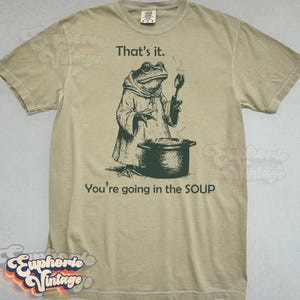 May include: Tan t-shirt with a frog illustration. The frog, wearing a robe, holds a spoon over a pot. Text above reads "That's it." and below, "You're going in the SOUP."