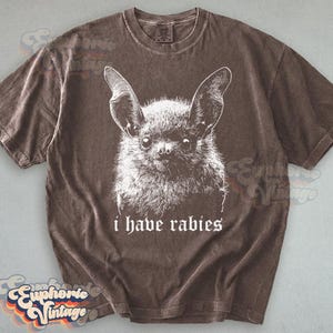 May include: Brown t-shirt featuring a white illustration of a bat with large ears and the text "i have rabies" in a gothic font. The shirt has a vintage, worn look.