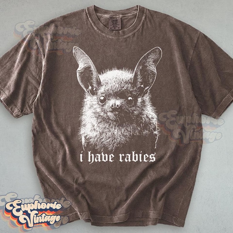 May include: Brown t-shirt featuring a white illustration of a bat with large ears and the text "i have rabies" in a gothic font. The shirt has a vintage, worn look.