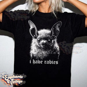 May include: Black t-shirt featuring a detailed, grayscale illustration of a bat's face. The bat has large ears and a furry appearance. Below the image, the text reads "i have rabies".