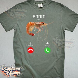 Op de afbeelding: Saliegroen t-shirt met een afbeelding van een garnaal en het woord "shrimp" erboven. De afbeelding lijkt op een telefoonscherm met "calling...", een rode weigerknop en een groene acceptatieknop.