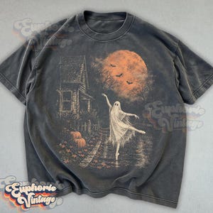 May include: A charcoal gray t-shirt featuring a graphic print of a ghost dancing in front of a house at night. The scene includes a large orange moon, flying bats, and pumpkins on the ground. The design has a distressed, vintage-inspired look.