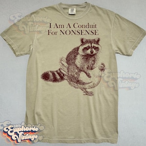May include: Tan t-shirt with the text "I Am A Conduit For Nonsense" above a graphic of a raccoon riding a chicken. The raccoon is brown and white, and the chicken is beige.