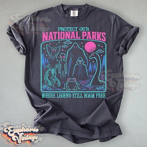 May include: A charcoal gray short-sleeve t-shirt featuring a graphic print with the text PROTECT OUR NATIONAL PARKS and WHERE LEGEND STILL ROAM FREE. The design includes a moth, alien, Bigfoot, vampire, frog, and sea monster in neon colors.