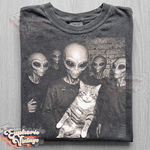 May include: Dark gray t-shirt featuring a group of aliens holding a cat. The graphic is in black and white, with the aliens in the center and a brick wall background. The shirt has a crew neck and the word "COMFORT" is printed on the collar.
