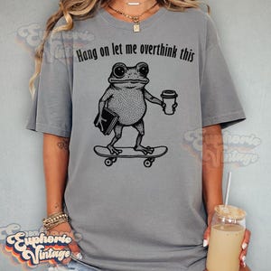 Puede incluir: Una camiseta gris de manga corta con un gr&aacute;fico negro de una rana de pie sobre un monopat&iacute;n mientras sostiene un libro y una taza de caf&eacute;. Sobre la rana, el texto dice Hang on let me overthink this, impreso en una fuente negra simple.