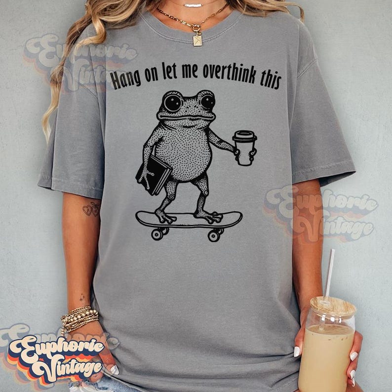 Puede incluir: Una camiseta gris de manga corta con un gr&aacute;fico negro de una rana de pie sobre un monopat&iacute;n mientras sostiene un libro y una taza de caf&eacute;. Sobre la rana, el texto dice Hang on let me overthink this, impreso en una fuente negra simple.