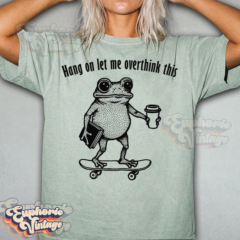 Puede incluir: Una camiseta de color verde salvia claro con un gr&aacute;fico negro de una rana sobre un monopat&iacute;n que sostiene un libro y una taza de caf&eacute;. El texto sobre la rana dice: "Hang on let me overthink this". Camiseta de manga corta y cuello redondo.