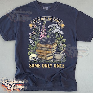 May include: A navy blue t-shirt featuring a graphic print of a black cat resting on a stack of books labeled Herbarium, Alchemia Herbalis, and Liber Mortis. The design includes a skull, a mortar and pestle, flowers, and the text All Plants Are Edible Some Only Once.