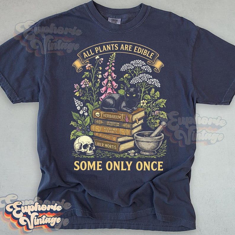 May include: A navy blue t-shirt featuring a graphic print of a black cat resting on a stack of books labeled Herbarium, Alchemia Herbalis, and Liber Mortis. The design includes a skull, a mortar and pestle, flowers, and the text All Plants Are Edible Some Only Once.