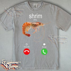 Op de afbeelding: Grijze t-shirt met een garnalenafbeelding. Het woord "shrimp" staat boven een realistische afbeelding van een gekookte garnaal. Daaronder staan twee telefoonpictogrammen met de woorden "Remind Me" en "Message".