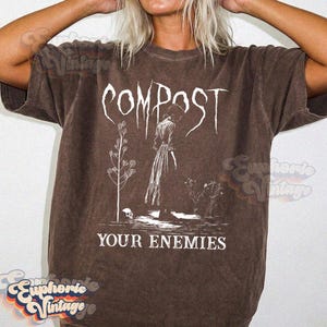 May include: A brown short-sleeve t-shirt featuring a white graphic print. The design includes the text COMPOST in a stylized font above an illustration of a person in a long dress standing near plants and a skull, with the text YOUR ENEMIES below.