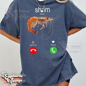 Op de afbeelding: Een marineblauw T-shirt met een garnalenafbeelding en de tekst "shrimp calling...". Het ontwerp bevat een telefooninterface met de knoppen "Remind Me" en "Message". De garnaal is oranje en wit.