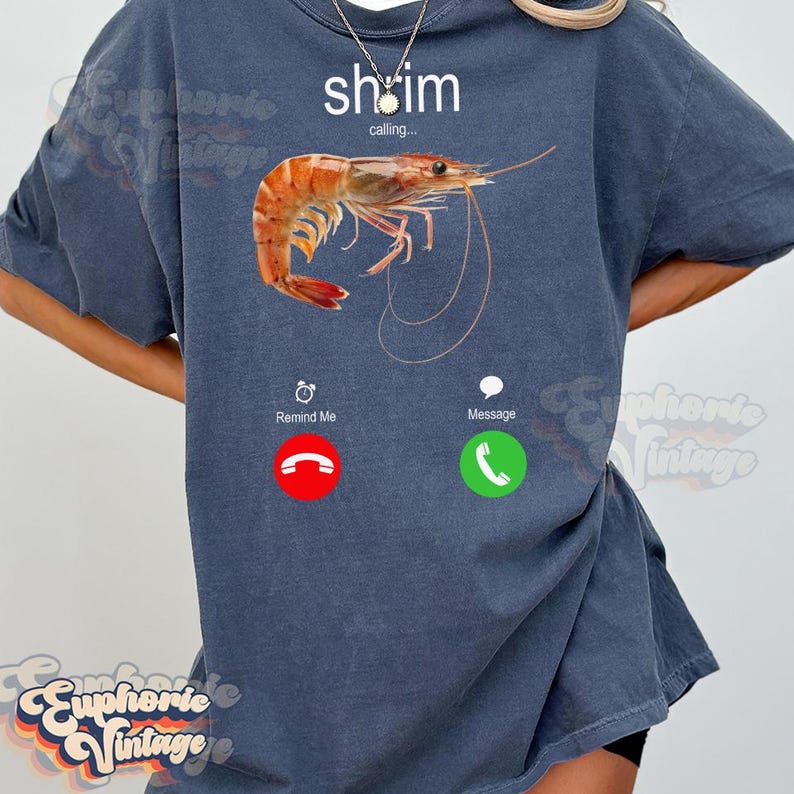 Op de afbeelding: Een marineblauw T-shirt met een garnalenafbeelding en de tekst "shrimp calling...". Het ontwerp bevat een telefooninterface met de knoppen "Remind Me" en "Message". De garnaal is oranje en wit.