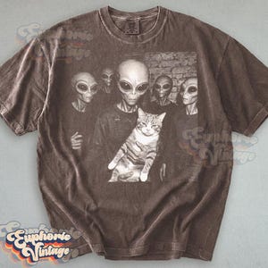 May include: Brown t-shirt featuring a graphic of several aliens with large eyes, one holding a tabby cat. The image has a vintage, distressed look. The shirt has a crew neck and short sleeves.