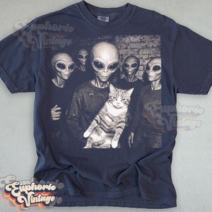 May include: Navy blue t-shirt featuring a black and white graphic of several aliens and a cat. The aliens have large black eyes and are holding the cat. The shirt has a vintage aesthetic.