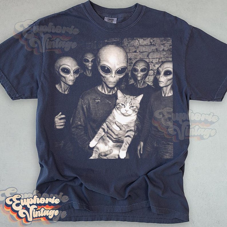 May include: Navy blue t-shirt featuring a black and white graphic of several aliens and a cat. The aliens have large black eyes and are holding the cat. The shirt has a vintage aesthetic.